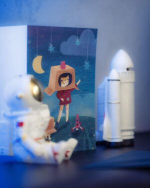 Biurko dziecka na którym stoi biała figurka kosmonauty, rakieta kosmiczna i granatowa kartka z dziewczynką bawiącą się w wyprawę w kosmos